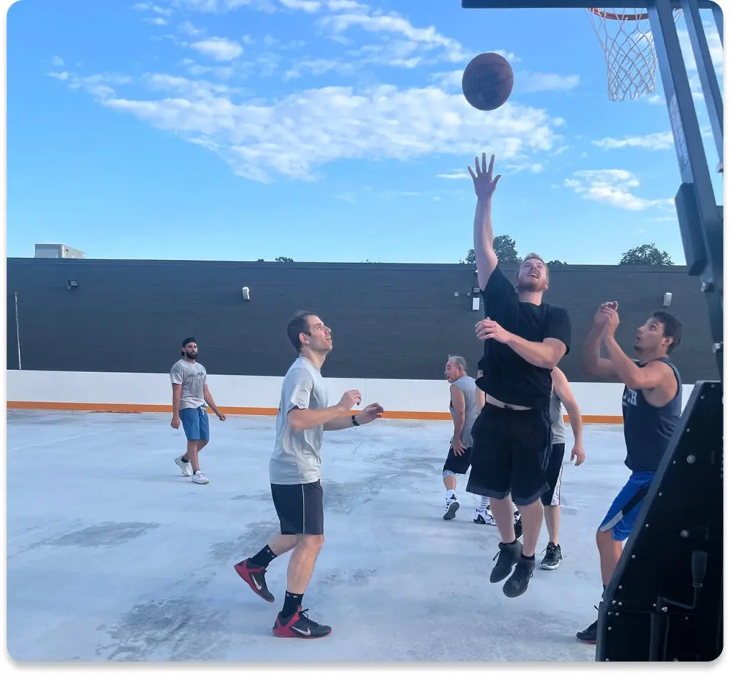 Six men play basketball on an outdoor court; one player in black jumps to shoot the ball towards the hoop while others watch or prepare to rebound.