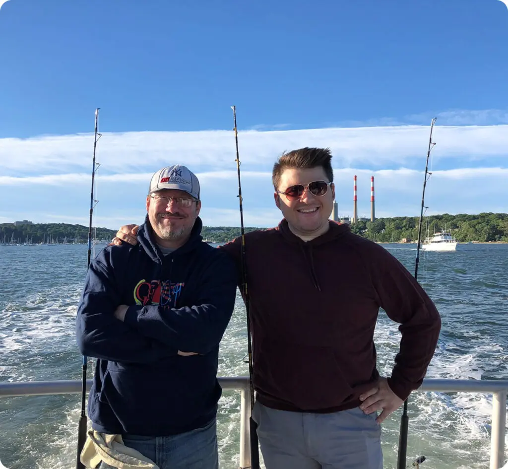 Two men stand smiling on a boat with fishing rods behind them, water and distant shoreline visible under a clear blue sky.