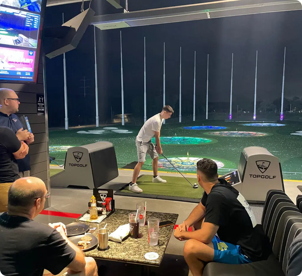 A man prepares to swing a golf club at a Topgolf driving range while three others watch and sit at a table with food and drinks.