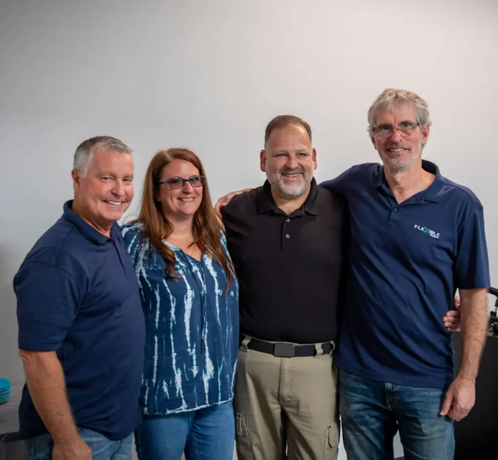 Four adults stand side by side, smiling at the camera against a plain background. Three men wear collared shirts and jeans; one woman wears a blue patterned shirt and glasses.
