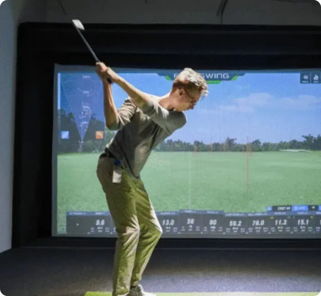 Person swinging a golf club indoors in front of a golf simulator screen displaying course graphics and data.