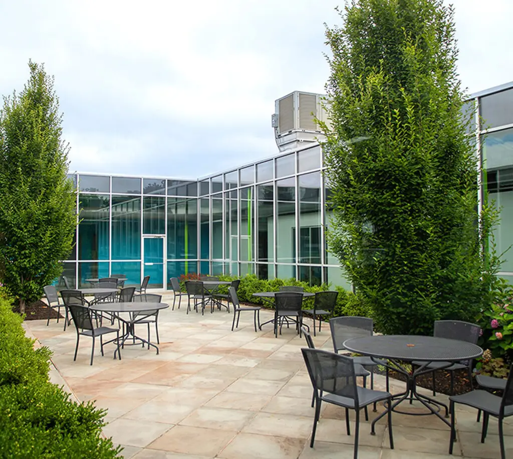 Outdoor patio area with metal tables and chairs, surrounded by greenery and trees, adjacent to a modern glass building under a cloudy sky.