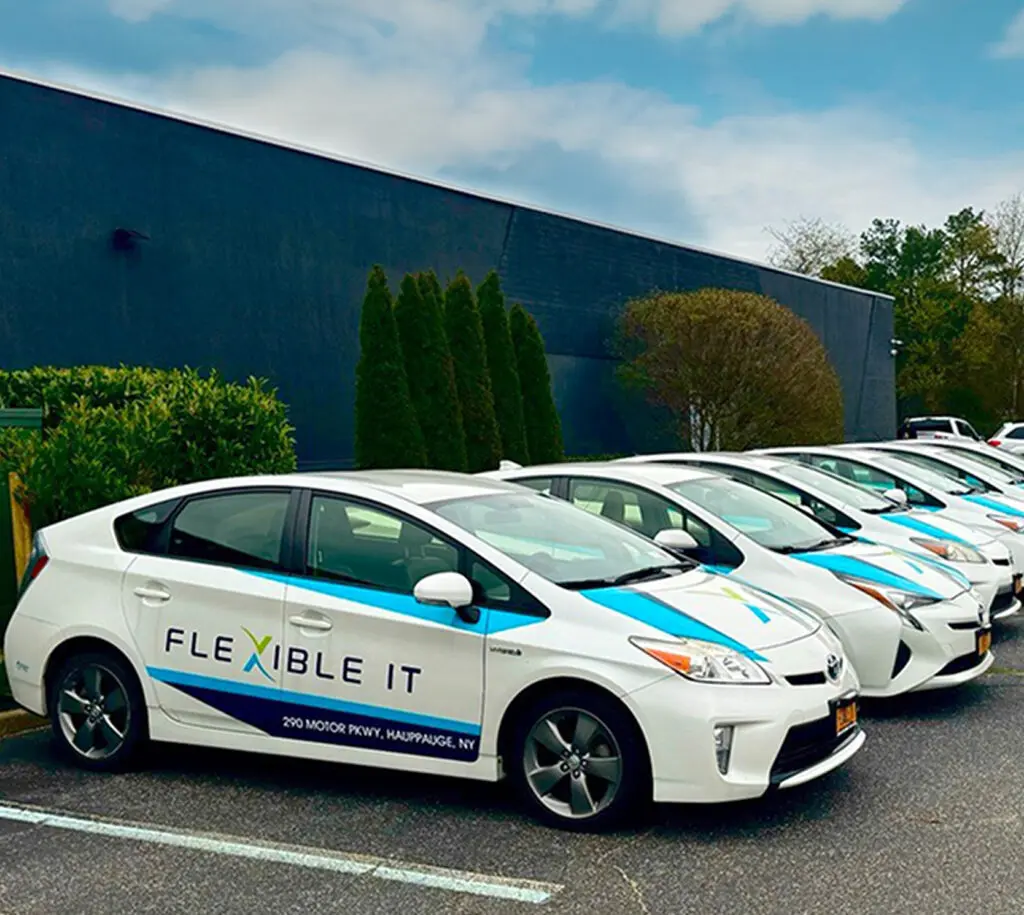 Several white sedans with "FLEXIBLE IT" branding are parked in a row outside a large industrial-style building on a cloudy day.