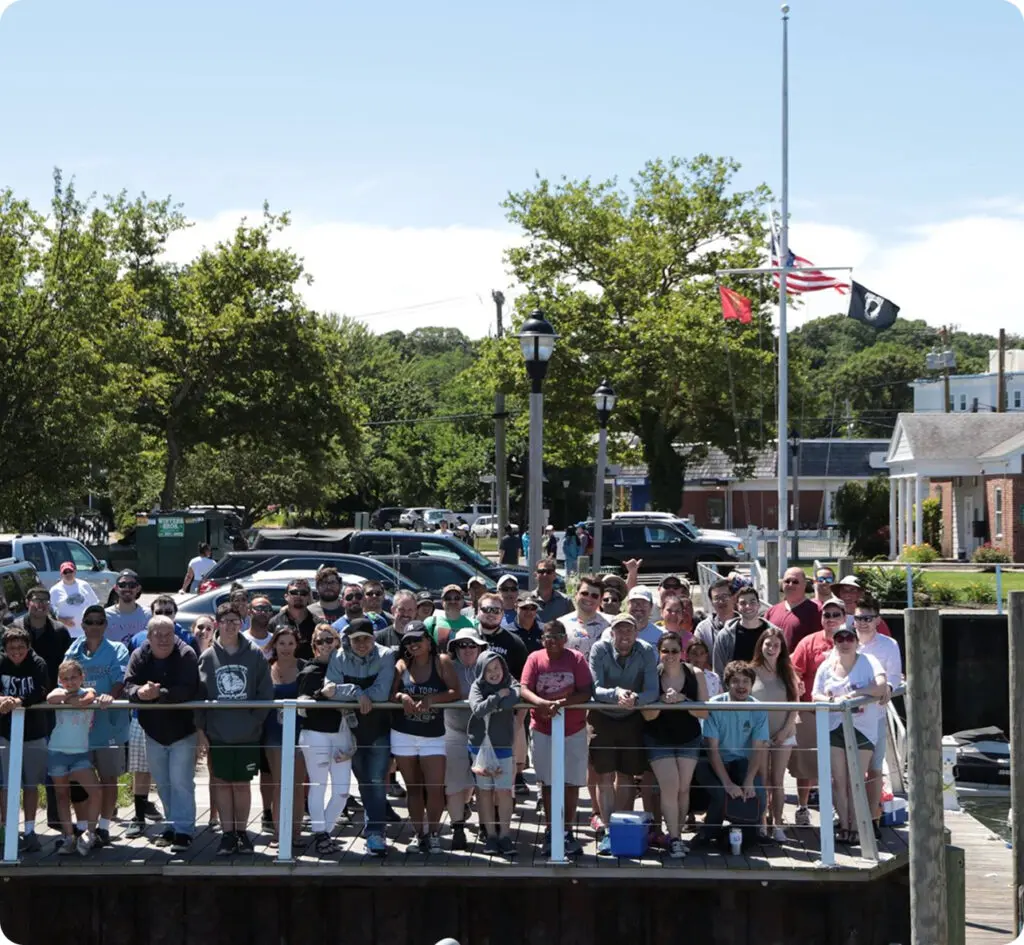 A large group of people stand closely together on a wooden dock, posing for a photo. Trees, parked cars, and buildings are visible in the background, with flags at half-staff.