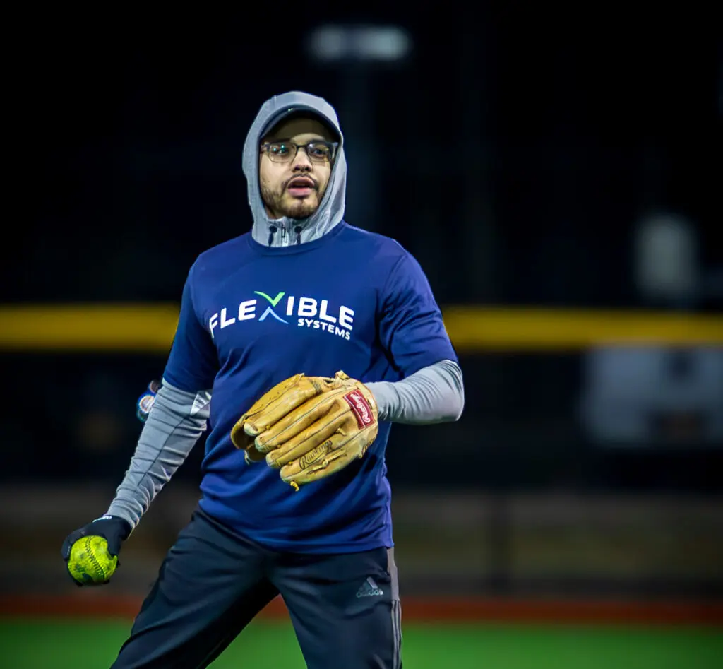 A person wearing a "Flexible Systems" shirt, hoodie, and glasses holds a softball and glove on a field at night.