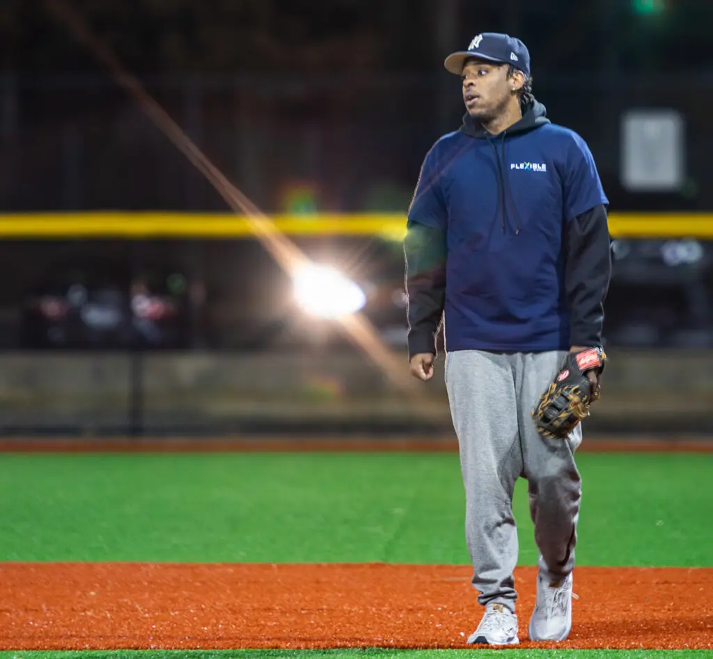 A man wearing a blue hoodie, gray sweatpants, and a baseball glove stands on an artificial turf field at night, with a bright light and cars in the background.