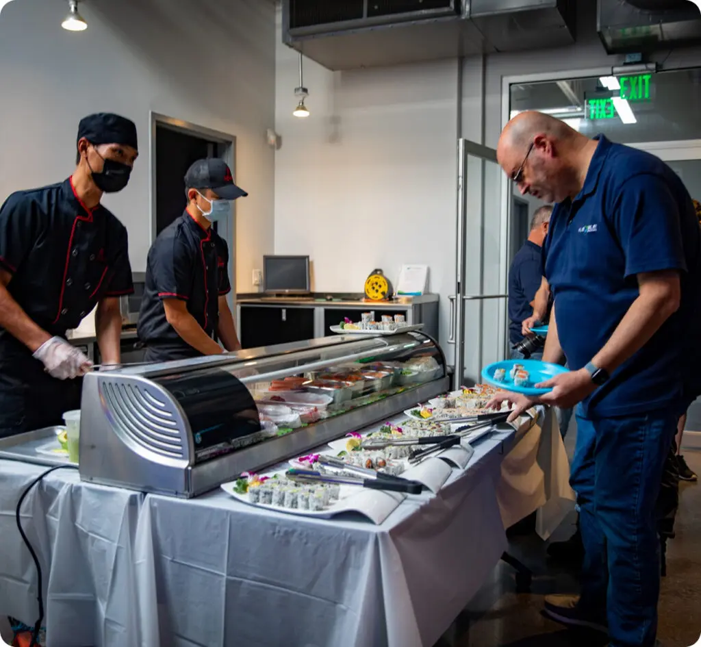 A man selects sushi from a buffet table staffed by two chefs wearing black uniforms and face masks in a modern indoor setting.