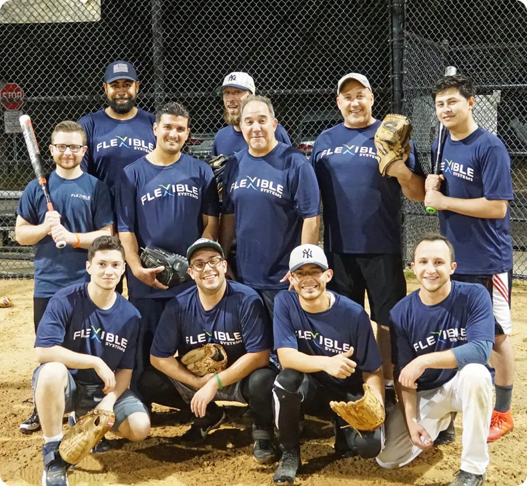 A group of eleven men in matching "Flexible" shirts pose together on a baseball field at night, holding bats and gloves.