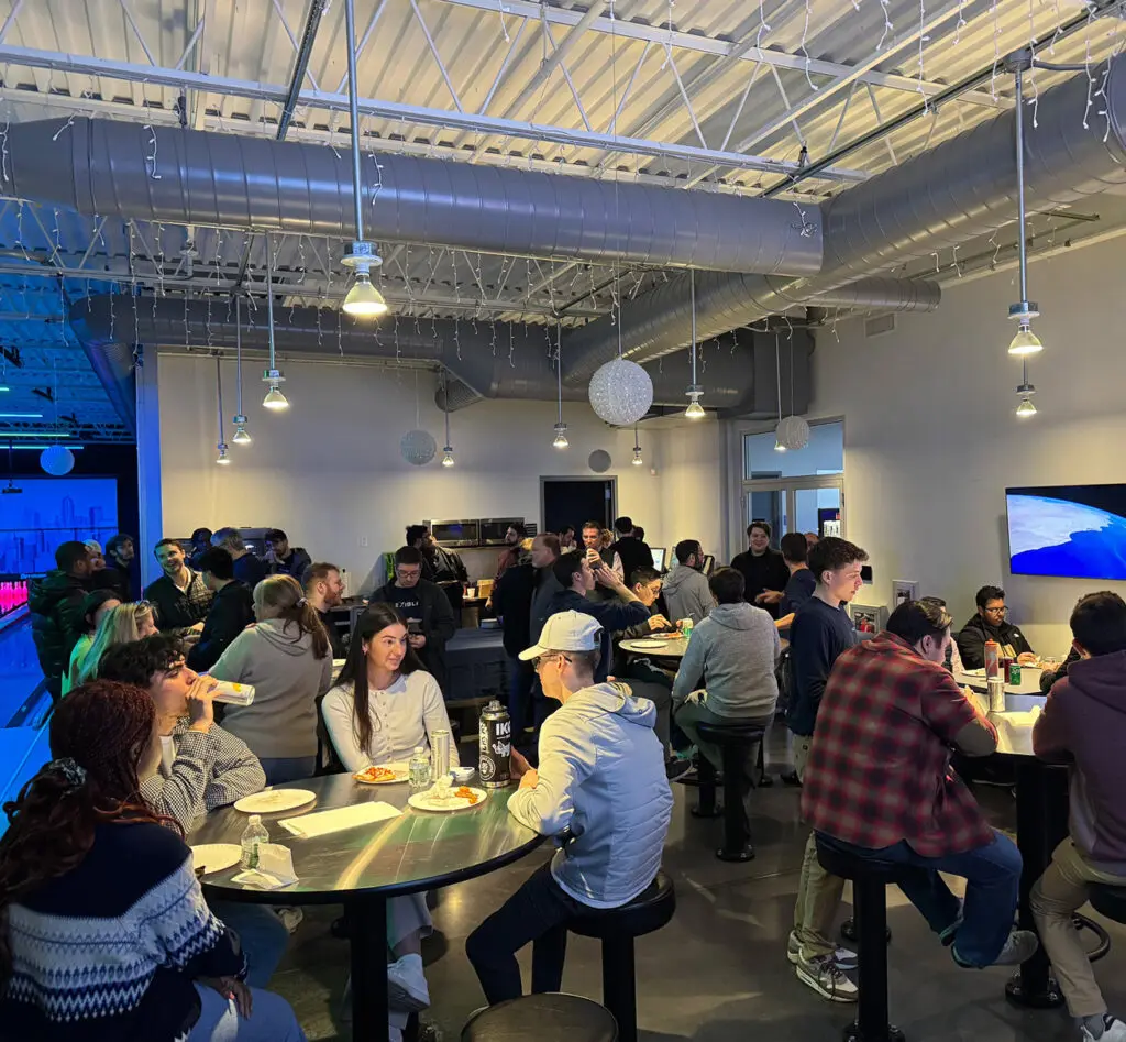 A group of people sit and stand around tables in a modern indoor space, eating, talking, and socializing under exposed industrial ceilings and hanging lights.