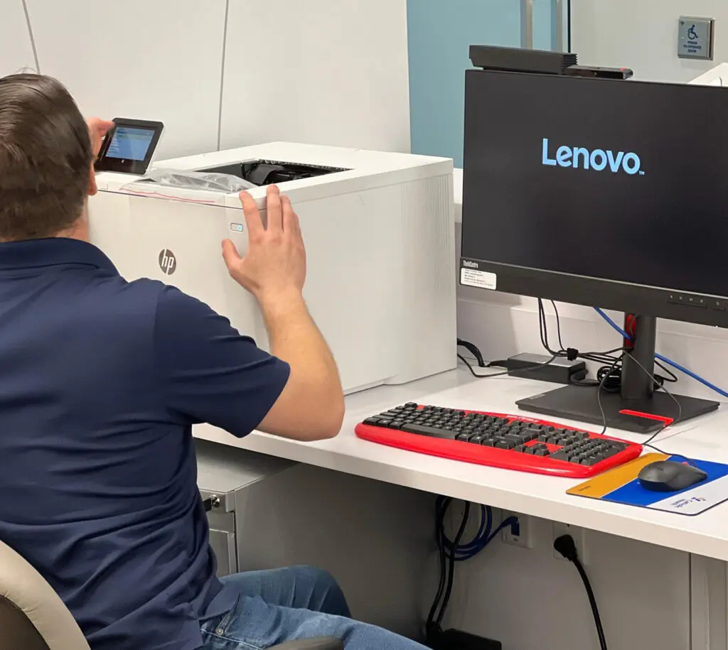 A person scans a barcode on an HP printer with a mobile device at a desk with a Lenovo monitor, red and black keyboard, and mouse.