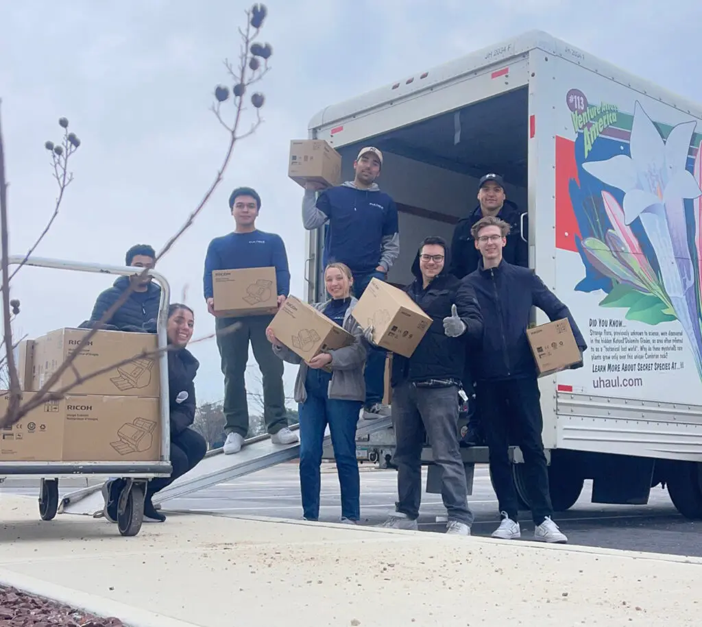 A group of people pose with boxes while unloading a moving truck parked on a curb. Some are holding boxes and others stand beside a loaded cart.