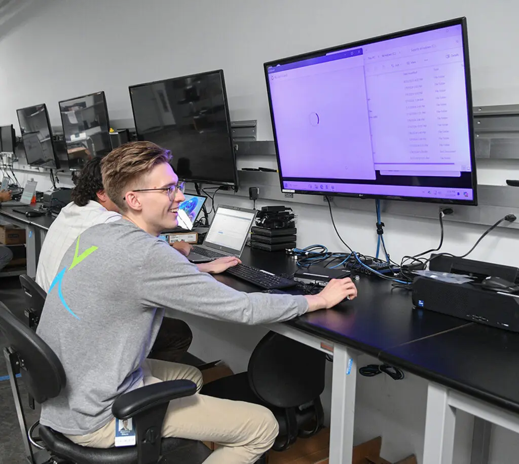 Two people sit at a desk with multiple monitors, one smiling at a large screen displaying a computer interface, working in a modern office or lab environment.