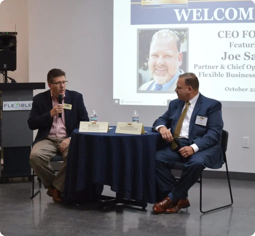 Two men in suits sit at a small table with microphones, participating in a CEO forum event with a presentation screen and speaker podium in the background.