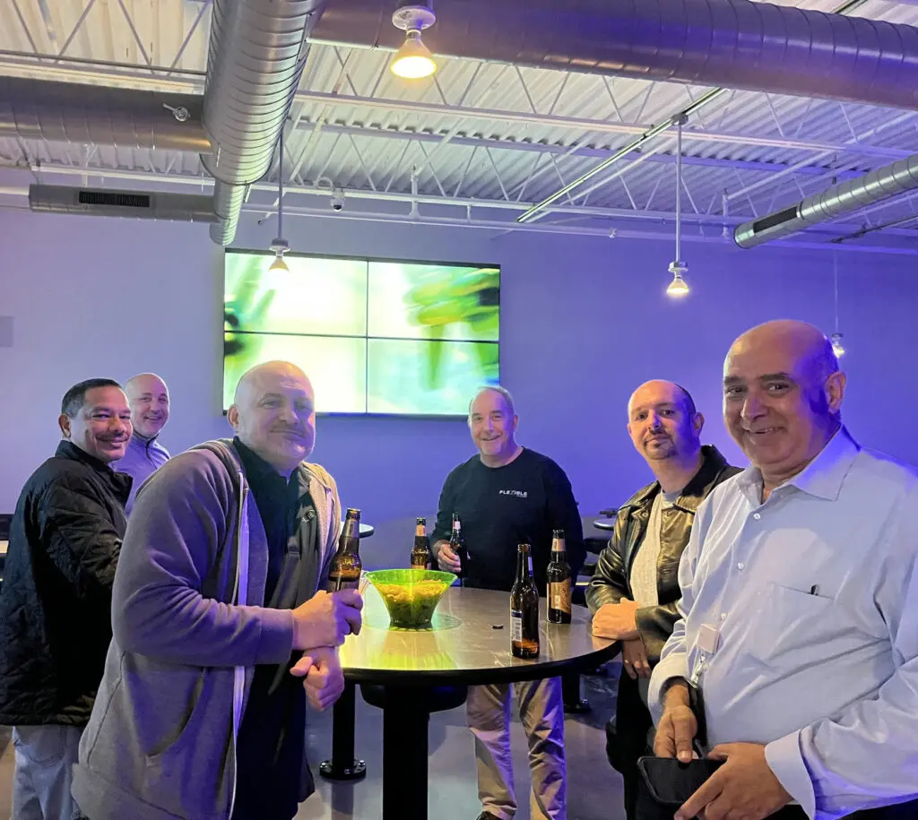 Six men stand around a tall table with drinks and snacks in a modern, industrial-style room with blue lighting.