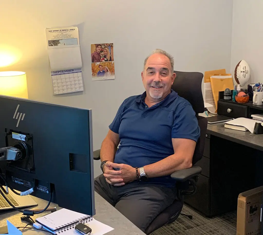 A man sits at a desk in an office, smiling at the camera. There is a computer monitor, paperwork, and office supplies on the desk, with a calendar on the wall behind him.