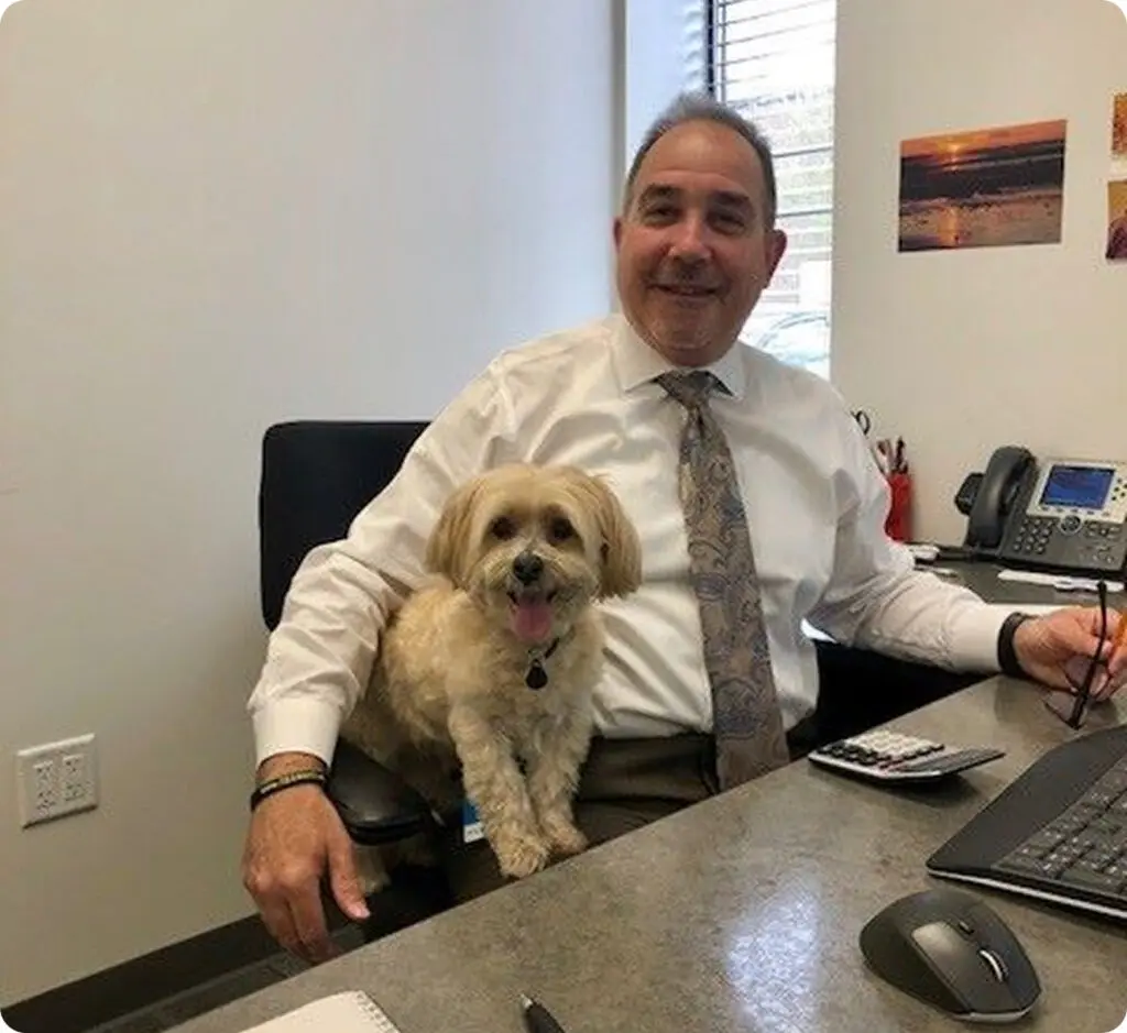 A man in business attire sits at a desk, smiling, with a small, light-colored dog on his lap in an office setting.