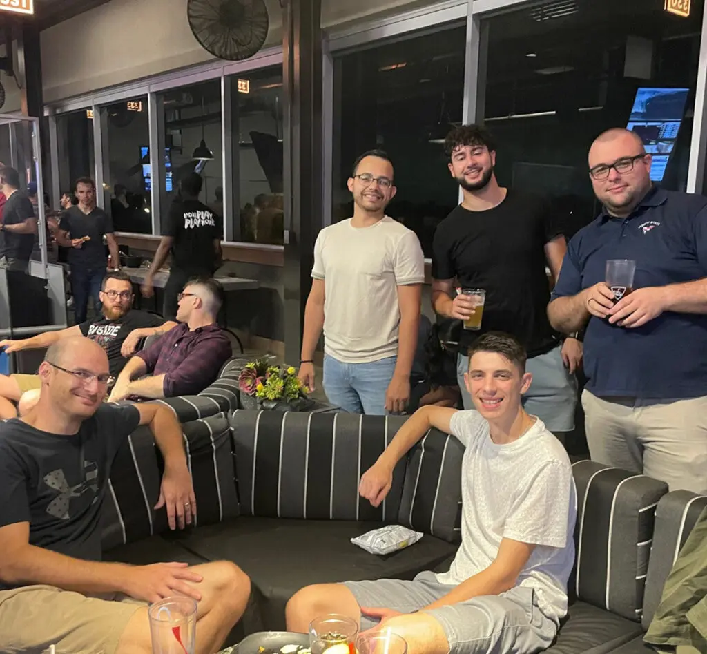 A group of men, some seated and some standing, pose together indoors on a striped sofa with drinks and snacks on the table in front of them. Other people can be seen in the background.