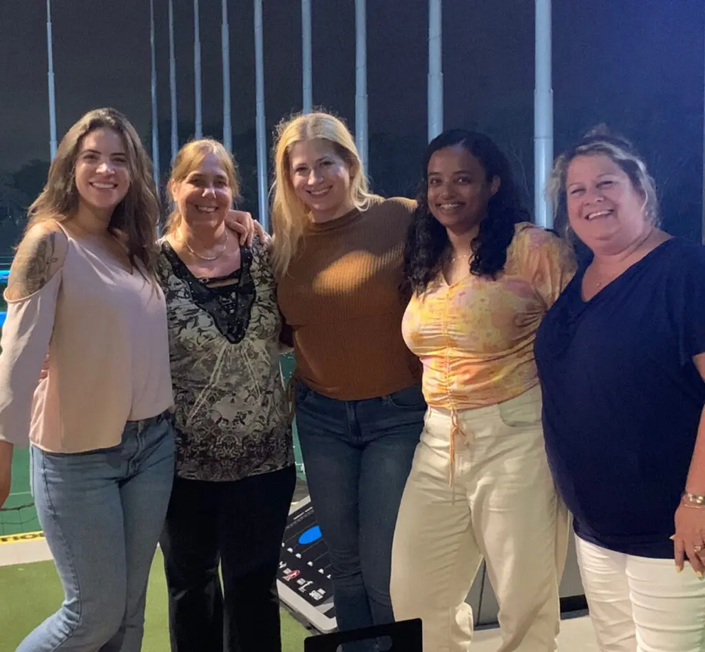 Five women stand together and smile for a group photo at an indoor golf driving range, posing in casual attire with golf nets visible in the background.