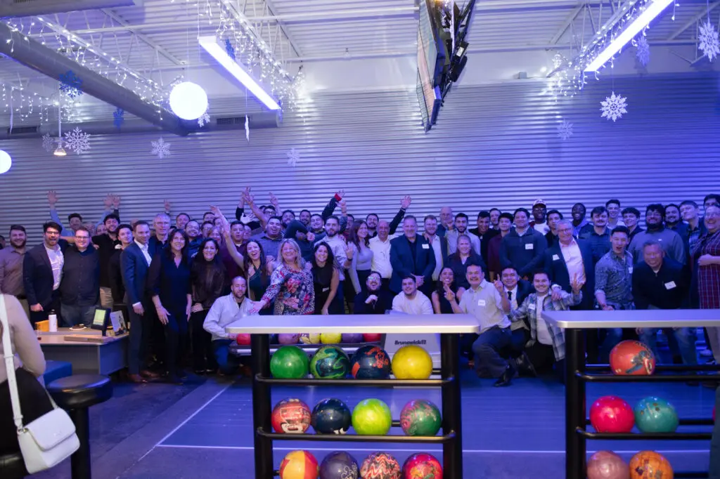 A large group of people pose together at an indoor bowling alley with colorful bowling balls visible in the foreground.