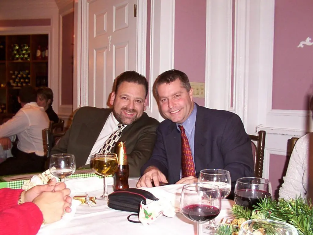 Two men in suits sit side by side at a restaurant table with drinks, smiling at the camera. Other people are seated nearby and holiday decorations are visible.