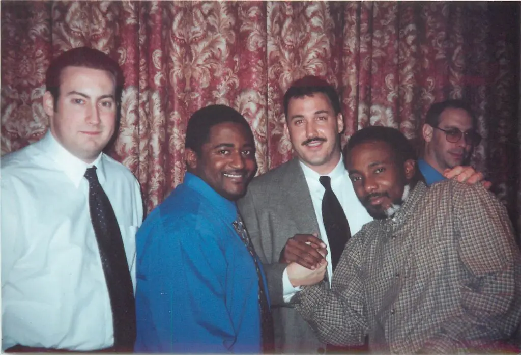 Five men in business attire pose together and smile in front of a patterned curtain, with two of them clasping hands in the foreground.