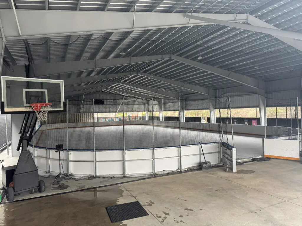 An indoor sports facility with a basketball hoop in the foreground and a fenced hockey rink in the background, under a metal roof.