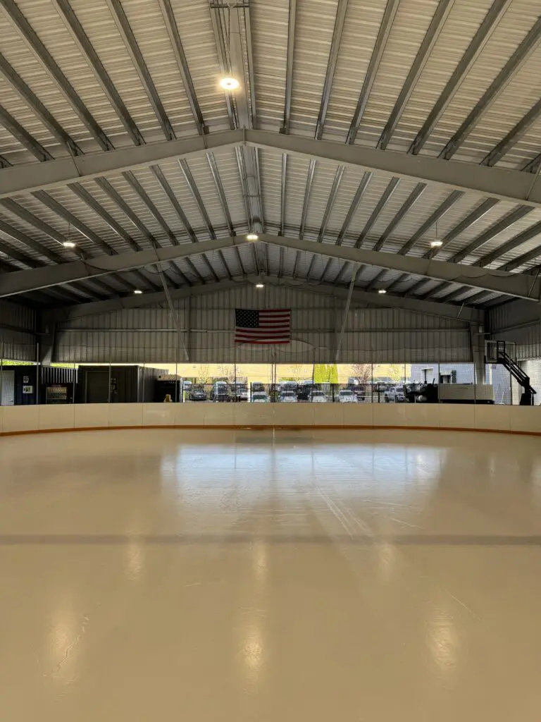 Empty indoor ice rink with a smooth surface, overhead lights on, and an American flag hanging on the far wall above large windows showing parked cars outside.