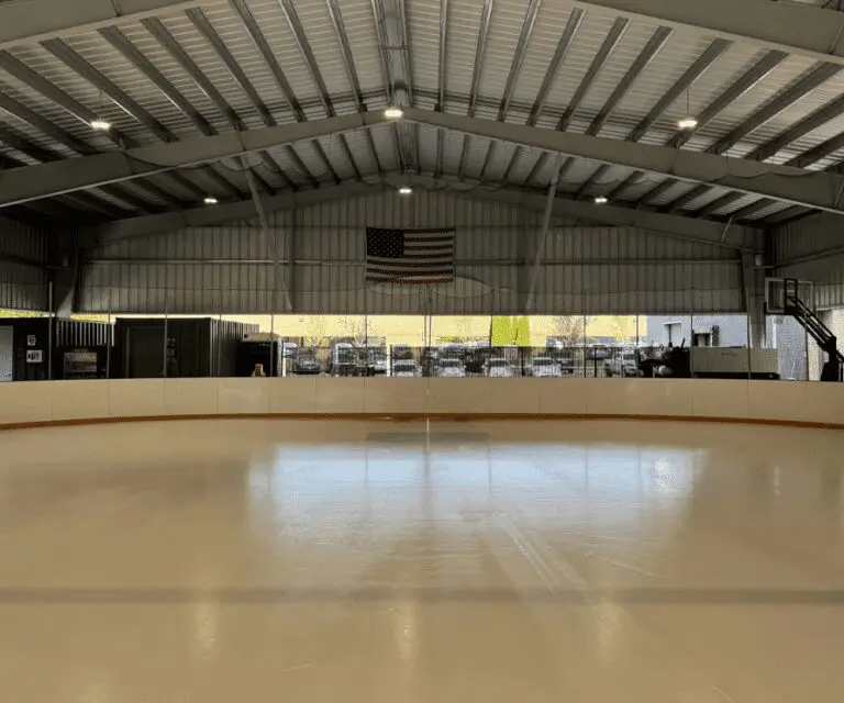 Indoor ice skating rink with smooth, empty ice surface, an American flag hanging from the ceiling, and large windows showing cars parked outside.
