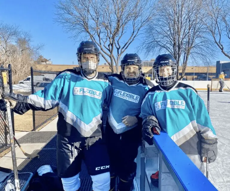 Three hockey players in teal jerseys and black helmets stand together, smiling, on an outdoor ice rink with trees and buildings in the background.