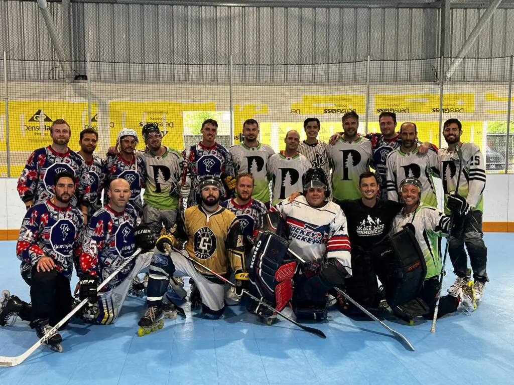 A group of men in hockey gear pose for a team photo on an indoor rink, with some wearing white jerseys and others in red and blue jerseys.