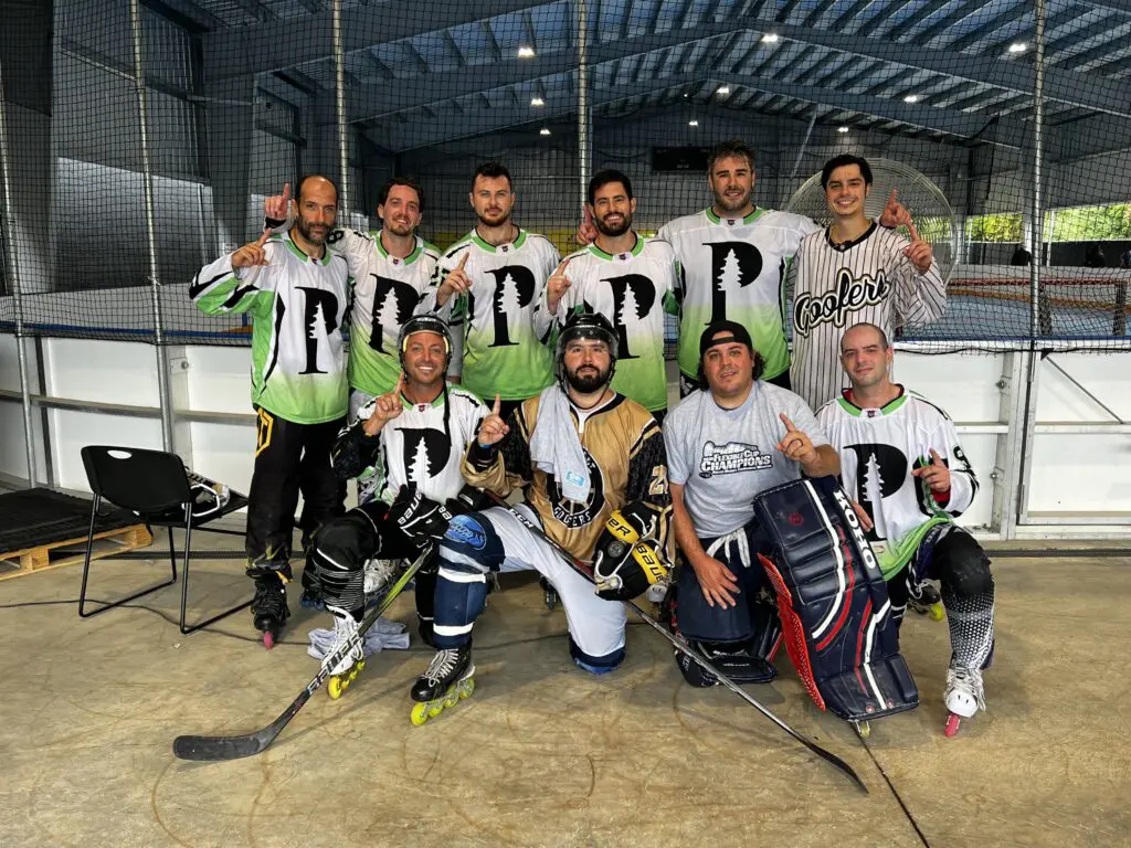 A group of nine adult men in roller hockey uniforms pose together on an indoor rink, some kneeling in front, all making a number one gesture.