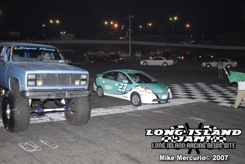 A lifted blue truck and a teal race car are side by side at the starting line of a racetrack at night, with a flagman waving a green flag nearby.