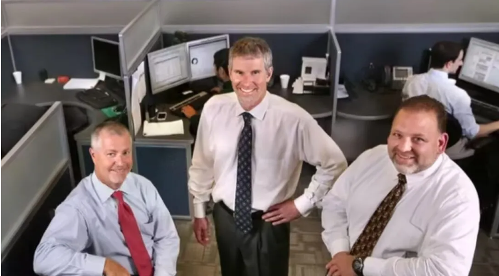 Three men in business attire stand and smile in an office cubicle area with computers and workstations in the background.