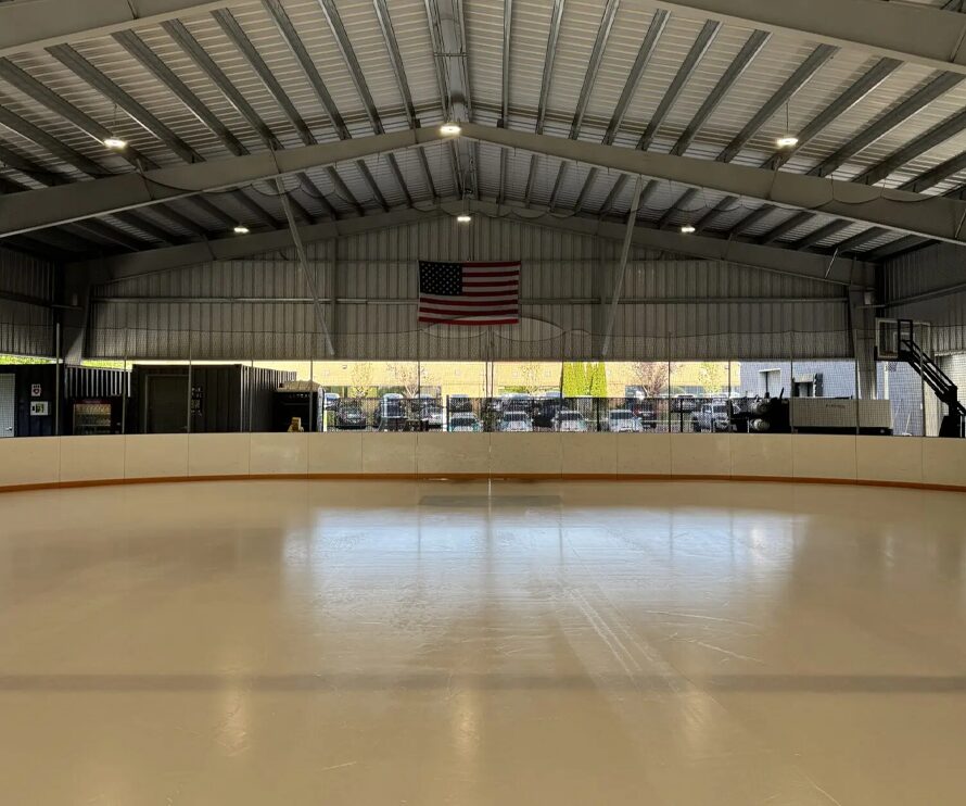 Indoor ice skating rink with smooth, empty ice surface, an American flag hanging from the ceiling, and large windows showing cars parked outside.