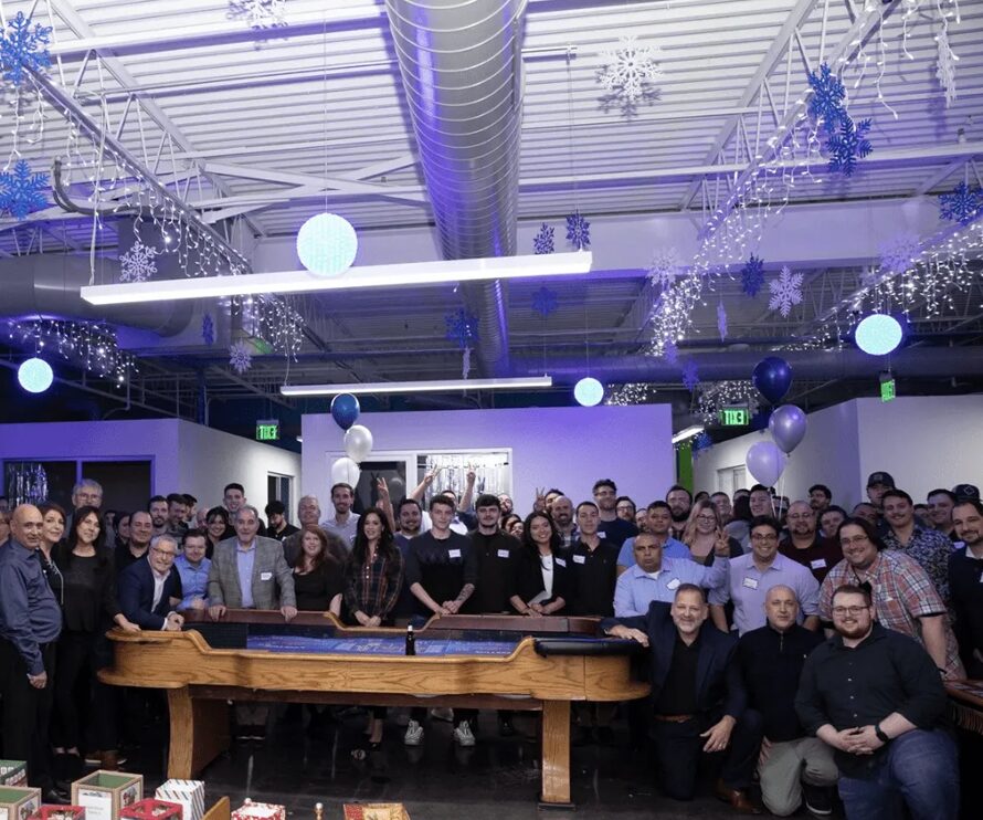 A large group of people pose together in an office space decorated with snowflakes, balloons, and string lights, standing behind a wooden shuffleboard table.
