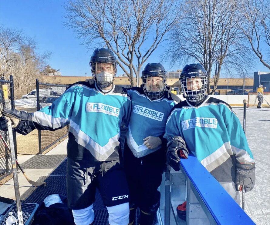Three hockey players in teal jerseys and black helmets stand together, smiling, on an outdoor ice rink with trees and buildings in the background.