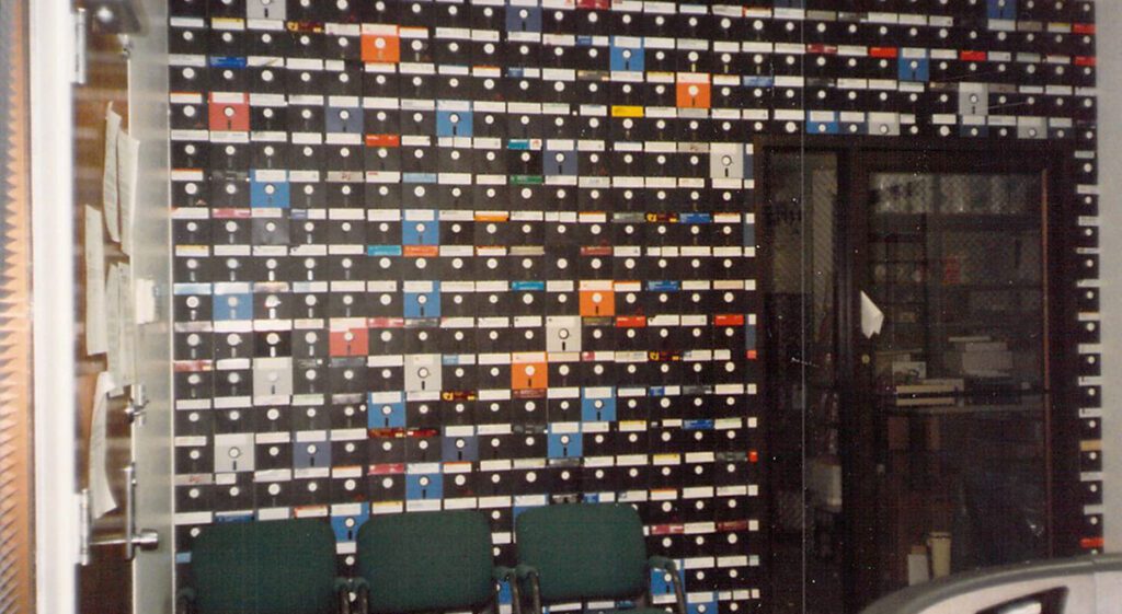 A wall filled with shelves holding numerous colorful floppy disks above three green chairs; a cabinet with glass doors is seen to the right.