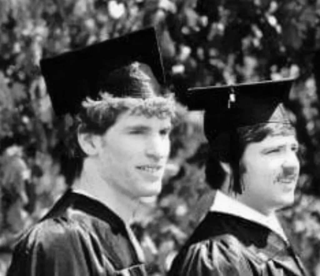 Two men wearing graduation caps and gowns stand outdoors, facing slightly to the right. The background is blurred with foliage.