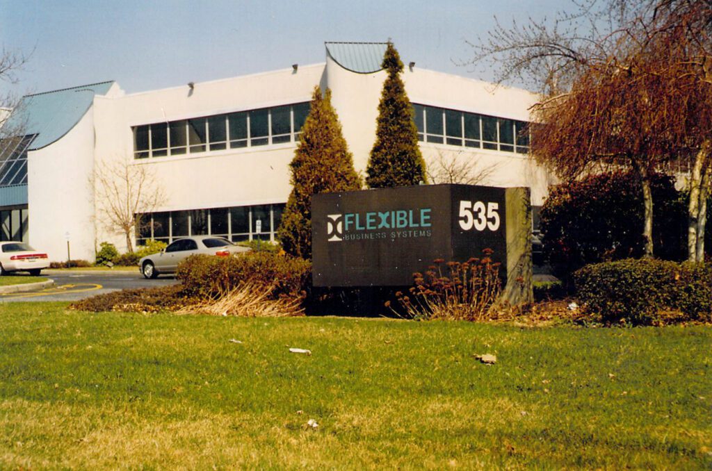 A business building with a sign in front reading "Flexible Business Systems 535," surrounded by trimmed bushes, trees, and a grassy lawn.