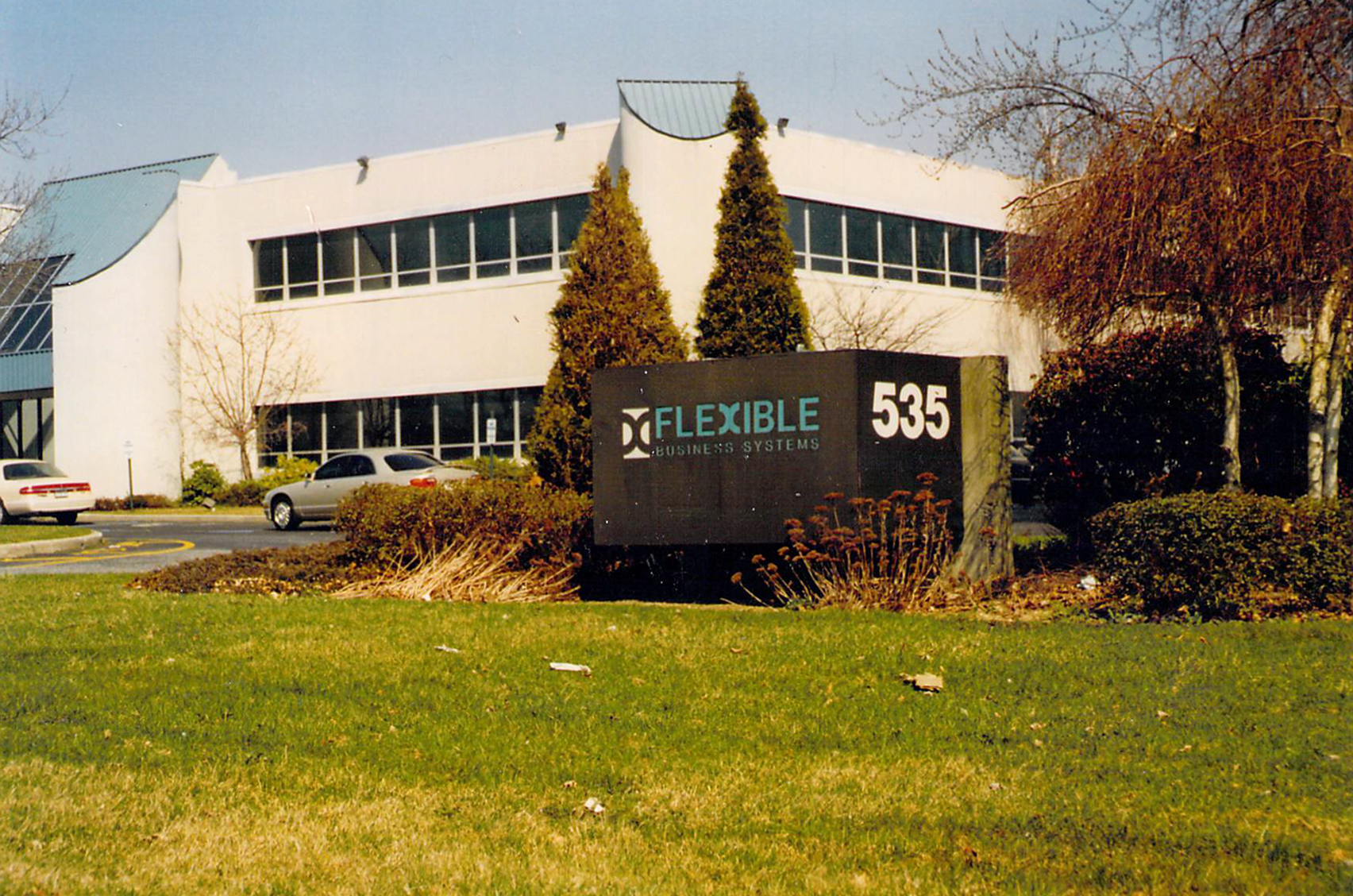 A business building with a sign in front reading "Flexible Business Systems 535," surrounded by trimmed bushes, trees, and a grassy lawn.