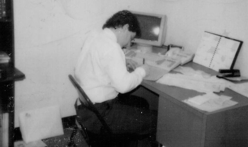 A person sits at a desk working on paperwork, with an old computer monitor, keyboard, and open planner nearby. The setting appears to be an office in the past.