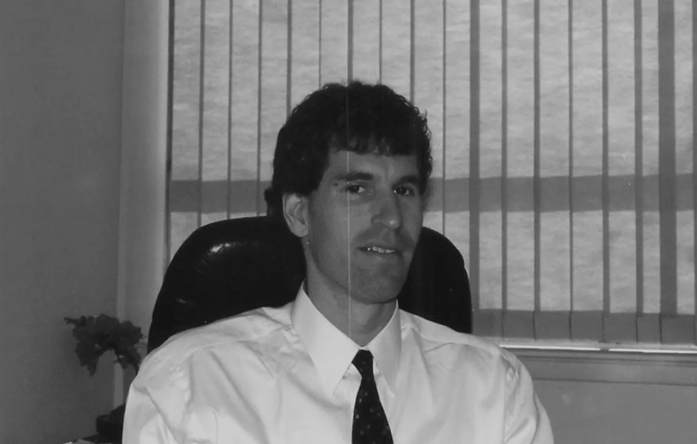 A man in a white shirt and tie sits in an office chair in front of a window with vertical blinds.