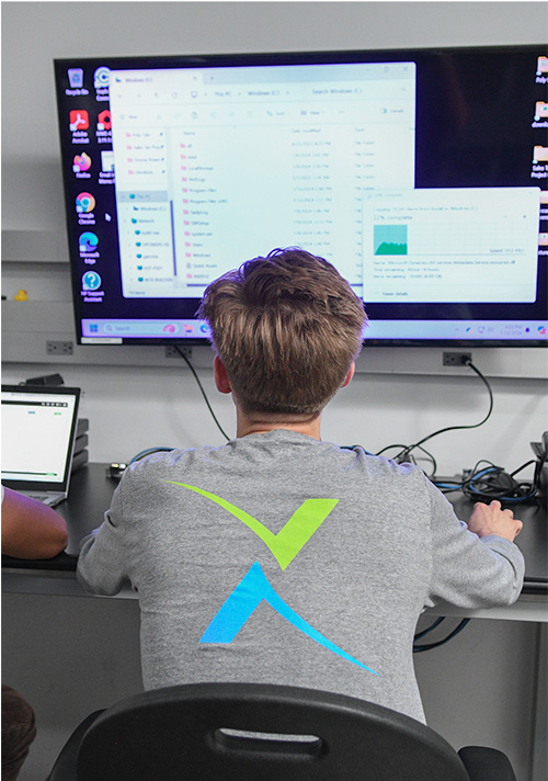 A person with light brown hair sits at a desk facing a large monitor displaying folders and graphs, embracing flexible IT solutions, with a laptop and cables neatly arranged on the desk.