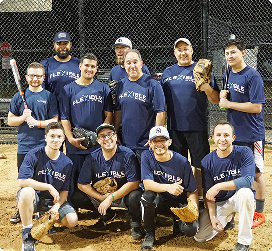 A group of eleven men wearing "FLEXIBLE" team shirts pose together on a baseball field at night, with gloves and a bat, showing what teamwork is about—flexible in spirit and strong in unity.