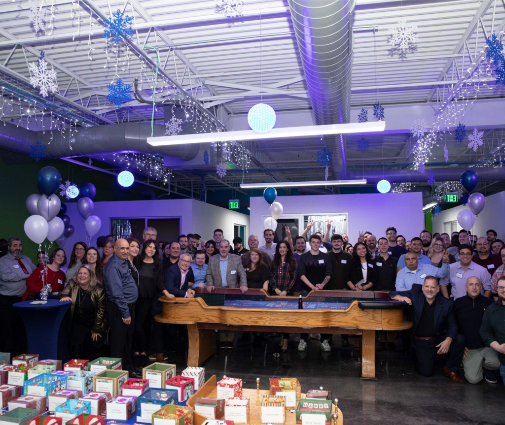 A large group of people pose for a photo at an indoor event with winter decorations, balloons, and tables of gift boxes in the foreground, highlighting the spirit of togetherness and celebrating the success of About Flexible IT.