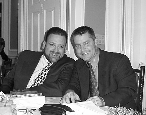 Two men in suits sit next to each other at a table, smiling at the camera in a formal indoor setting, discussing about flexible IT solutions.